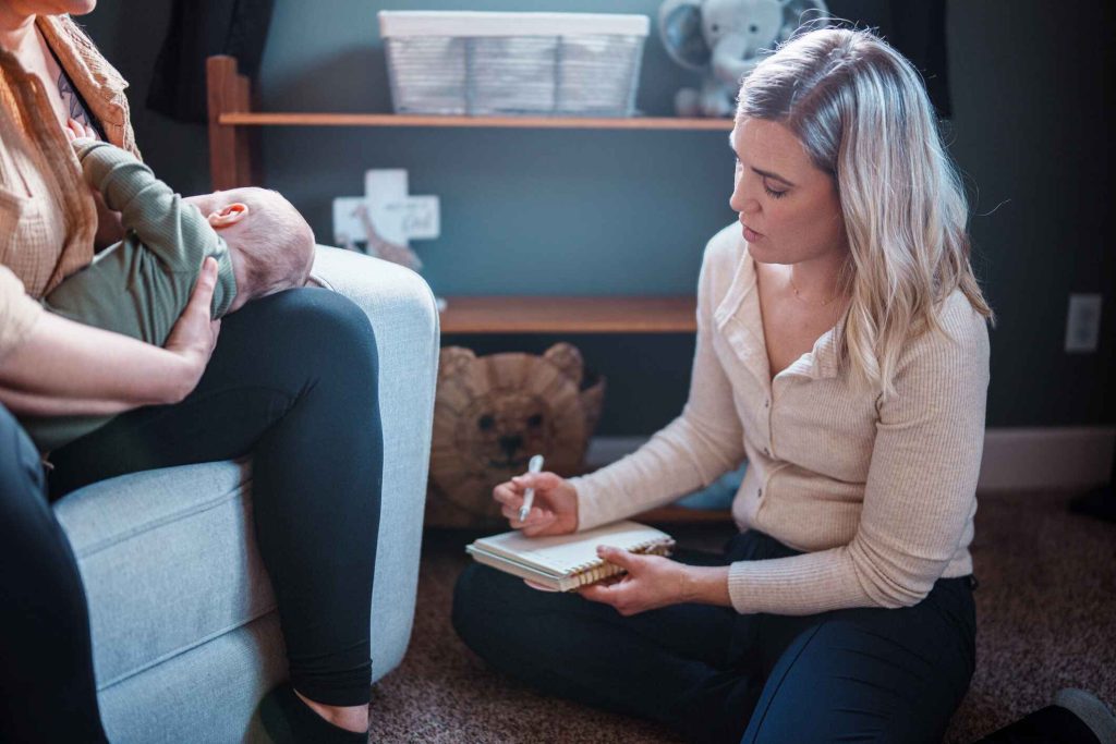 Mother holding a baby during a professional supervised visitation session, with a facilitator taking notes in a supportive environment.