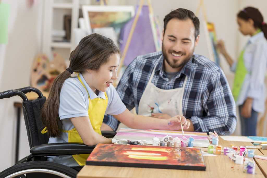 Girl in wheelchair painting with instructor in art class, fostering creativity and expression for children with special needs.