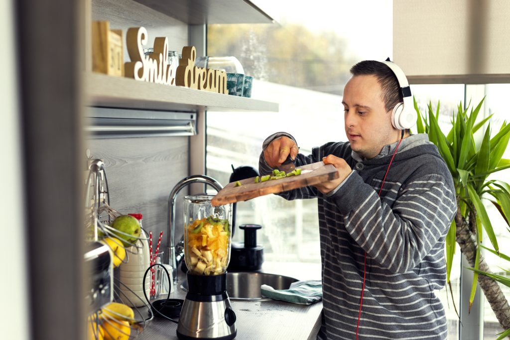 Person with a disability preparing food in a modern kitchen, wearing headphones, with a blender and fresh fruits visible, illustrating Supported Independent Living and personal empowerment.