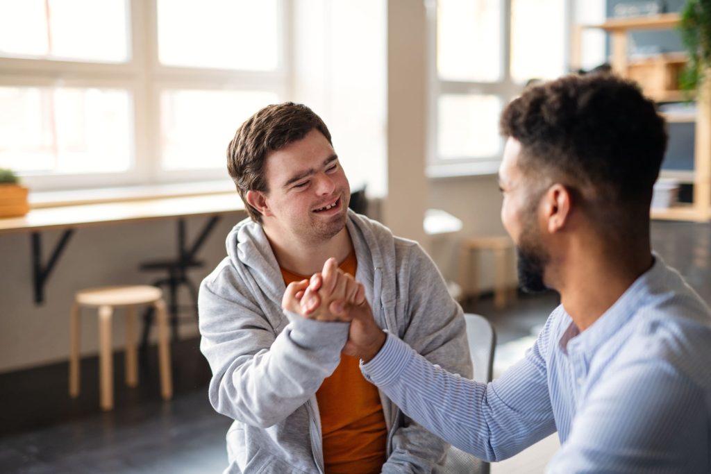 Two men engaging in a supportive interaction, smiling and holding hands, in a bright, modern indoor setting, representing the importance of connection and empowerment in NDIS Supported Independent Living contexts.