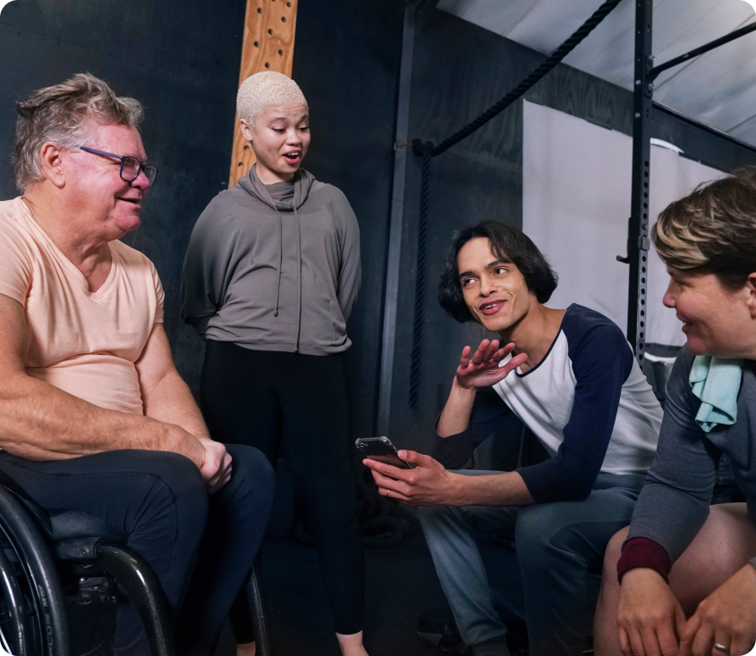 Group of four diverse individuals engaging in conversation and sharing a smartphone, illustrating community support and social interaction in a relaxed environment, relevant to Ability to Achieve’s focus on social participation and wellbeing.