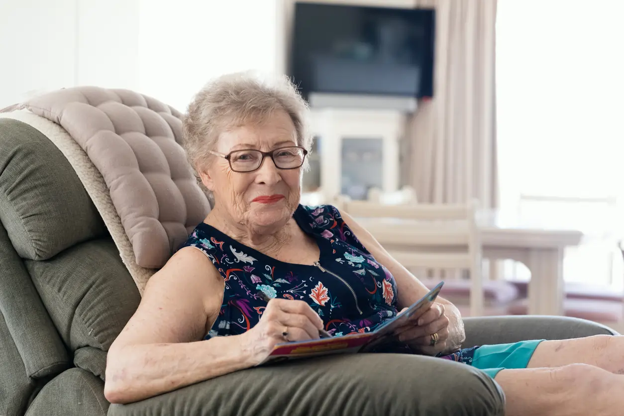 Older woman sitting comfortably in a chair, reading a book, in a bright living room, representing NDIS disability support services and tailored care for seniors.