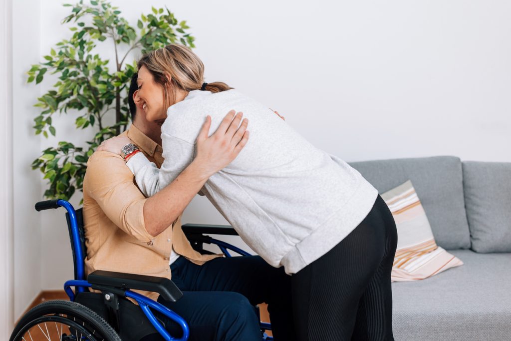 Woman embracing a man in a wheelchair, expressing support and connection, in a bright indoor setting with a plant and sofa, illustrating the importance of companionship in disability support services.