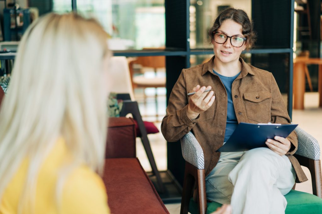 Woman in a brown jacket holding a clipboard, engaged in a conversation with another person, in a modern setting, highlighting disability support services and empowerment.