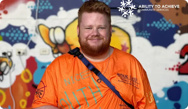 Smiling young man in an orange shirt, standing in front of a colourful mural, representing Ability to Achieve community services focused on disability support and inclusion in The Entrance.