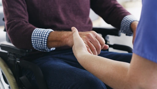 Hands of a healthcare professional holding the hand of a person in a wheelchair, symbolising support and care in the context of NDIS funding eligibility and assistance.
