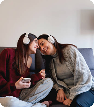 Two women wearing headphones, sitting closely together on a sofa, sharing a joyful moment while listening to music, reflecting connection and support in a cozy home environment.