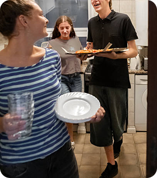 Group of young adults in a kitchen, smiling and engaging, with one person holding a plate and glass, another carrying food on a tray, reflecting community and support in a home-like environment.