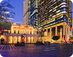 Historic building illuminated at dusk, surrounded by modern skyscrapers, representing the blend of heritage and contemporary architecture in Sydney's urban landscape.