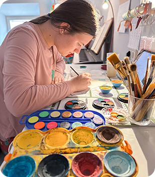Person engaged in painting with watercolours at a creative workspace, showcasing various paint palettes and brushes, reflecting a focus on artistic expression and personal development.