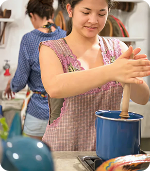 Young woman in a pink apron stirring a blue pot, demonstrating cooking skills in a home kitchen, with another person in the background, reflecting community and independence in a supportive environment.