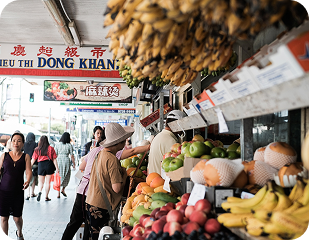 Busy street market scene featuring vendors selling fresh fruits and vegetables, with shoppers browsing and a sign reading "DONG KHANH" above.