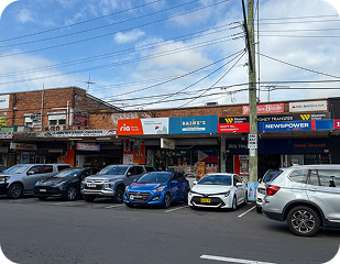 Street view of a commercial area in Sydney, featuring various shops and parked cars, representing community accessibility for disability services by Ability to Achieve.