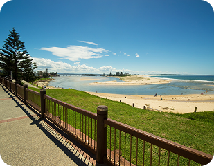 Scenic view of a beach and riverbank in a sunny location, featuring a wooden fence, green grass, and distant city skyline, relevant to Ability to Achieve's service areas in Sydney.