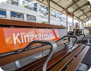 Kingsgrove train station bench with "Kingsgrove" signage, featuring a passenger waiting, highlighting accessibility in the area serviced by Ability to Achieve.