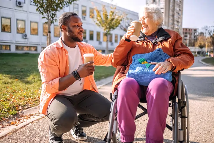 Support worker sharing coffee with elderly woman in wheelchair, enjoying community engagement outdoors, highlighting NDIS community participation and social inclusion.
