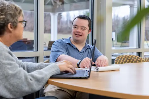 Person with Down syndrome engaging in conversation with a caregiver at a table, illustrating person-centred disability support services relevant to NDIS provider selection.