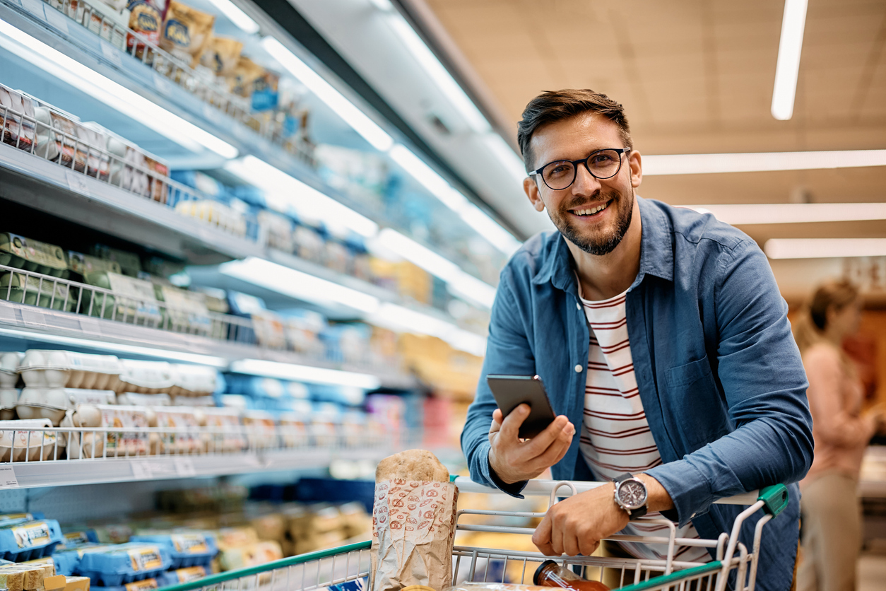 Man smiling in grocery store with shopping cart, holding smartphone, surrounded by shelves of food products, illustrating community engagement and independence in daily activities.