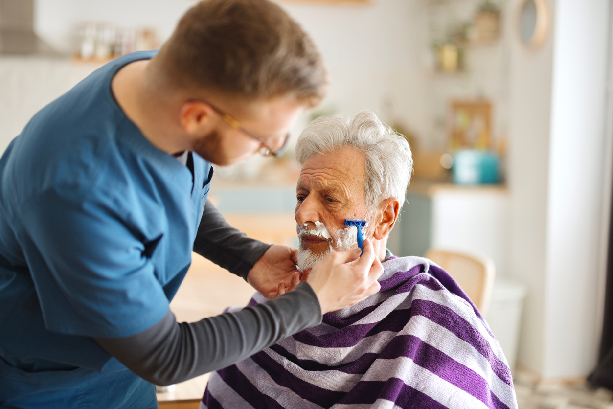 Caregiver assisting elderly man with personal grooming, shaving facial hair, emphasizing support in personal care activities for enhanced independence and self-esteem.