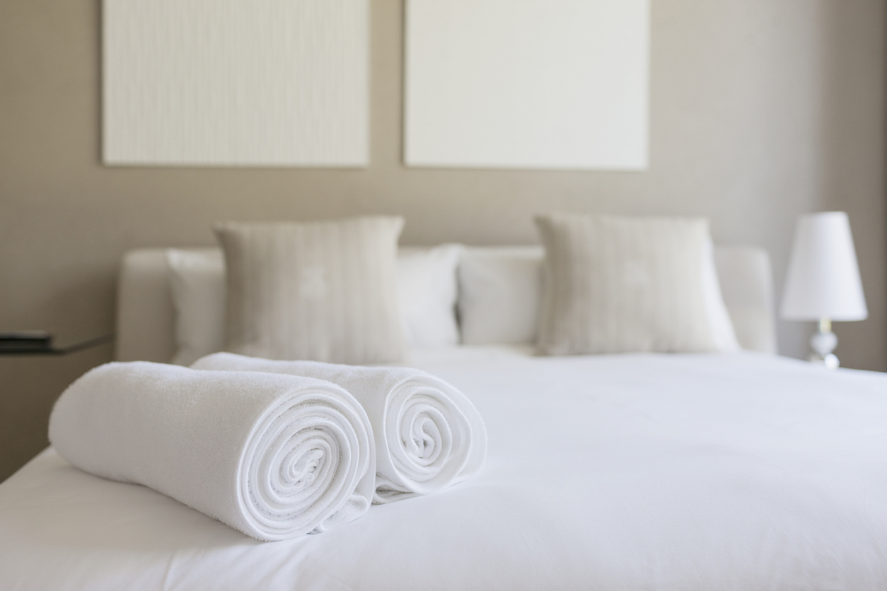 Cozy bedroom setting with neatly rolled white towels on a made bed, featuring soft pillows and a modern lamp, highlighting a welcoming atmosphere for supported accommodation.