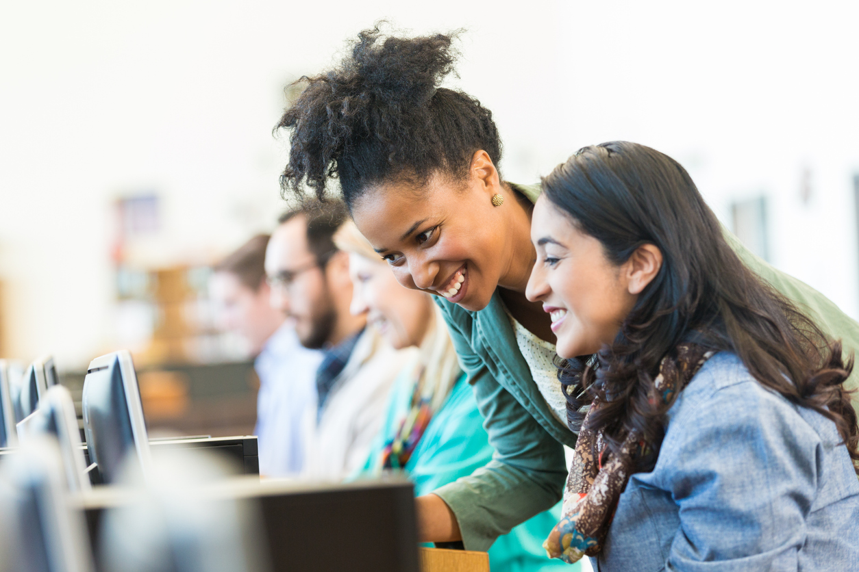 Two women engaged in a supportive learning environment, smiling while interacting with a computer, surrounded by others in a skills development setting.