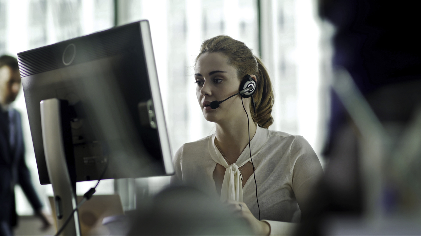 Woman wearing a headset, engaged in conversation at a computer, illustrating support services in a professional environment.
