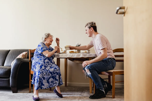 Elderly woman in blue floral dress engaging joyfully with a younger man at a table, illustrating supportive interaction in a home setting related to NDIS Supported Independent Living services.